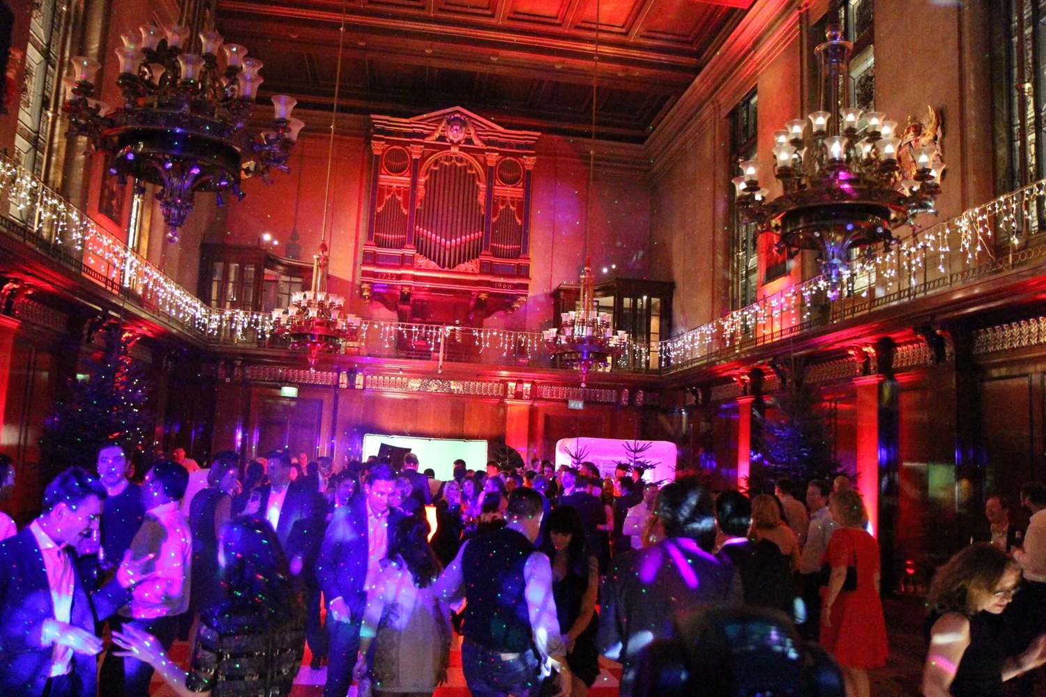 Guests dancing at a party in a grand chandeliered hall bathed in purple and red party lights