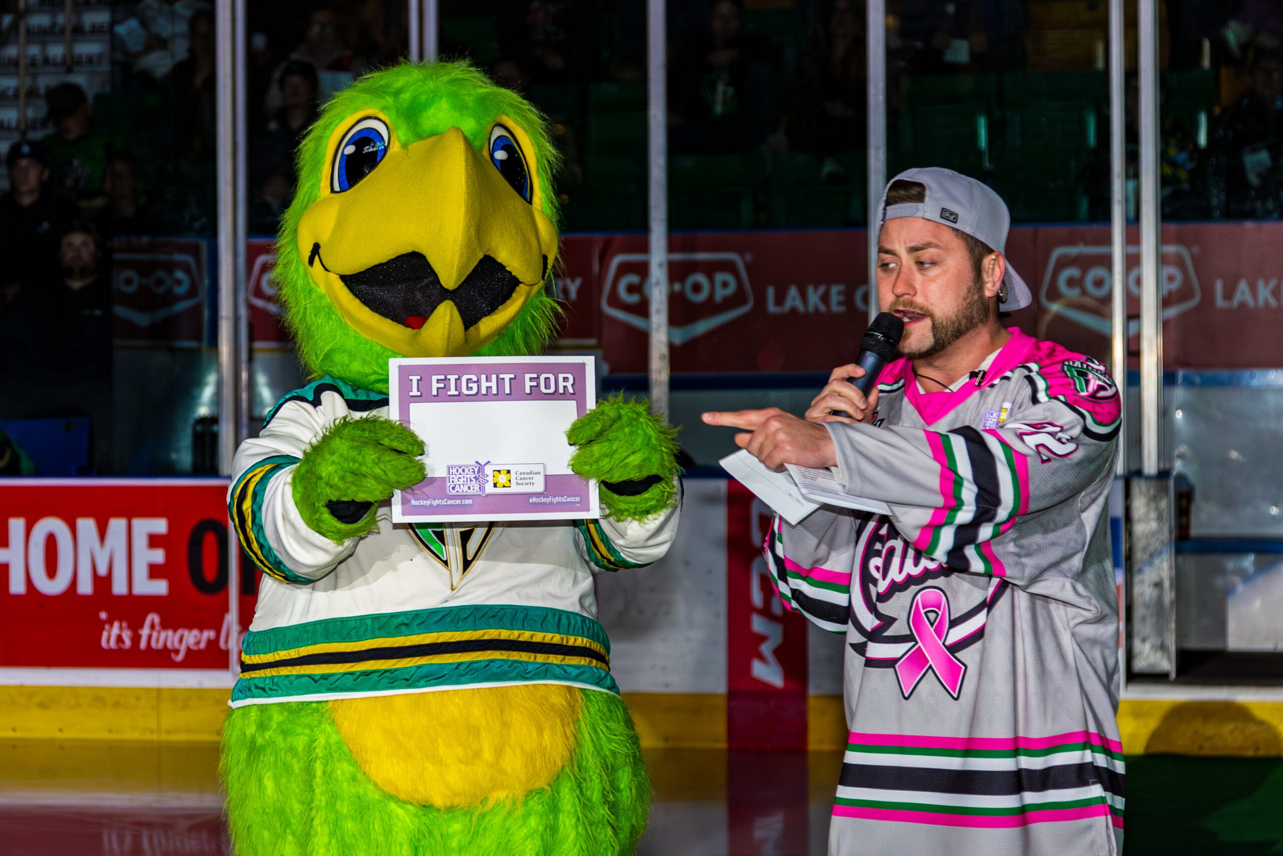 Tanner Fetch on the ice at the Art Hauser Centre in a pink-ribbon Raiders jersey, microphone in hand, pointing to the Raiders' green bird mascot holding a Hockey Fights Cancer 'I Fight For' sign