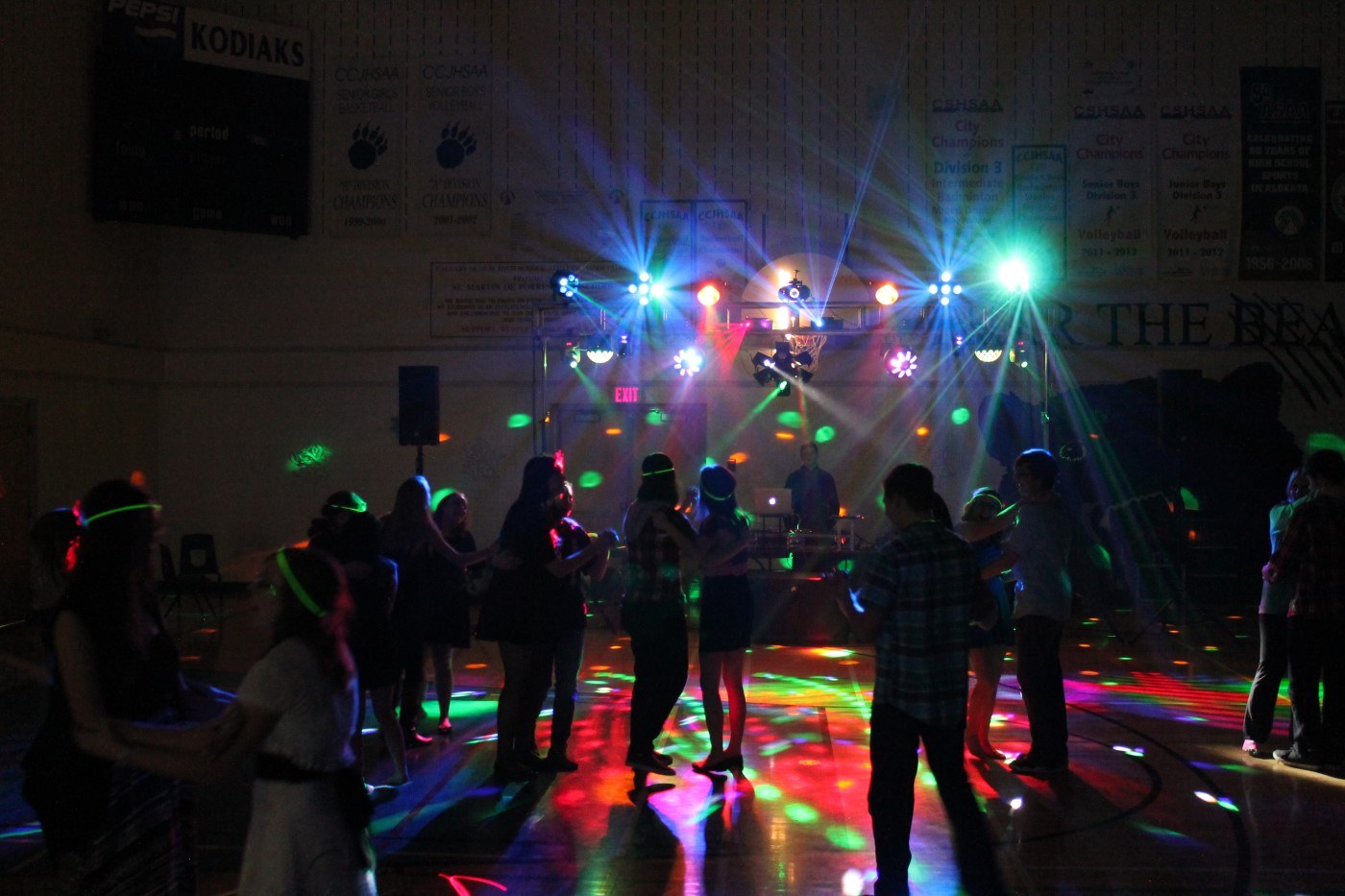Students slow-dancing at a real school dance in a gymnasium under colourful DJ light beams, the DJ booth at the far end beneath the Kodiaks scoreboard and championship banners
