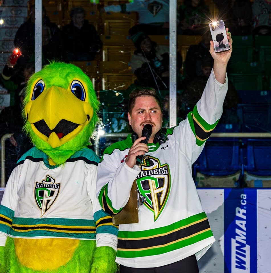 Tanner Fetch in his Prince Albert Raiders jersey, microphone in hand, hyping the crowd beside the Raiders' green mascot at the Art Hauser Centre